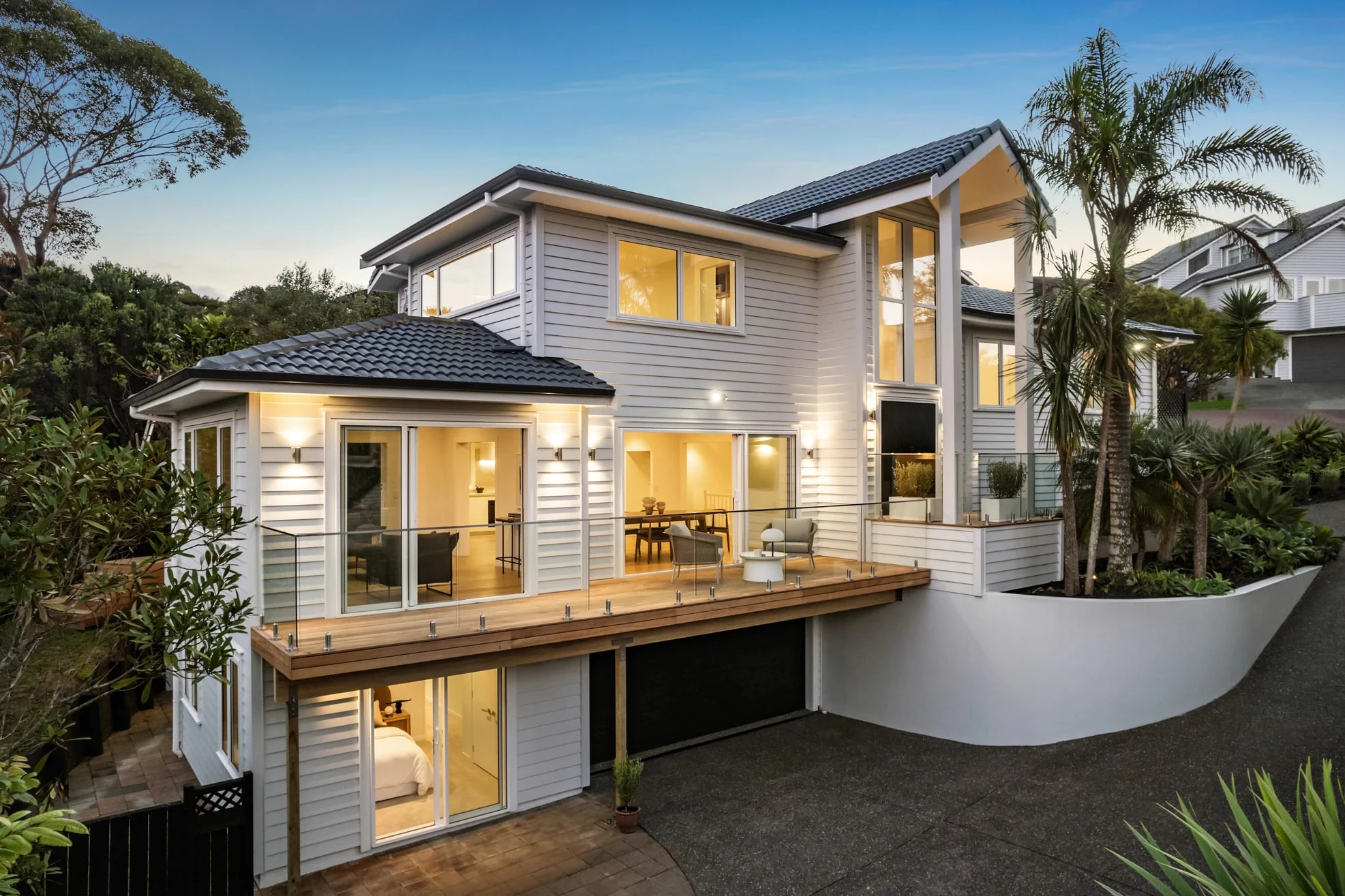 The recladded Campbells Bay home at dusk, white weatherboard over two levels with a glass-balustraded deck